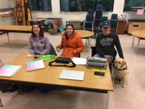 Jessica, Jocelyn, and Alex are sitting behind a table in a classroom. On the table are Braille books, a green cloth bag, a Brailler, an iPhone, a bag of cards, a BrailleNote in its case, 2 slates and styluses, a MacBook Pro, and Braille paper. Zandra is to the right of Alex.