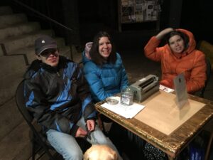 Alex, Jessica, and Jocelyn are sitting at a table outside the General Store. On the table are a Sign, a Perkins Brailler, a stack of tickets, a stack of bookmarks, and a donation jar.