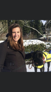 A photo of Shauna and guide dog, Abner, taken at the guide dogs for the blind Oregon campus, in Boring, Oregon in December 2016. It is a close-up photo of  Shauna  and Abner with snow in the background. Shauna has long brown hair, and is smiling, and is wearing a black winter jacket. Abner is wearing his bright yellow and black raincoat, GDB harness, collar and leash. Abner is a black Labrador retriever, and in this photo he is two years old.