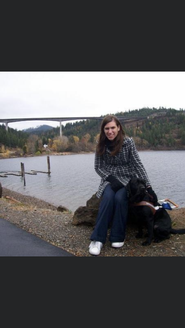 A photo of Shauna and Rafferty. Shauna  is wearing a dark plaid jacket, dark blue jeans, and white running shoes, and has long brown hair. Rafferty is a black lab retriever and is wearing his GDB harness, collar and leash. They are seated on a large rock at lake Coeur d’Alene in Idaho. There is a beautiful landscape behind them of the lake, and a bridge off in the far distance.