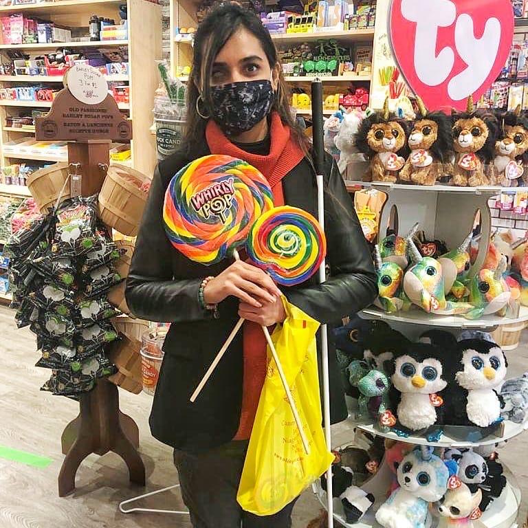 This photo shows Wajiha standing in a candy shop holding two huge lollipops across each other in her hands. The bigger lollipop is about 12 inches across and the smaller one is about 8 inches across. She’s looking at the camera, smiling and wearing a black face mask with tiny flowers patterned on it. She’s wearing a trendy pink cashmere sweater and a black blazer over it, that has leather sleeves while the rest of the fabric looks like cotton, with medium gray jogger pants and black flip-flops. Her waist length brown and blonde straight hair is down with a silky white and black knotted headband on top of her head with side bangs framing her face. Her long white cane is resting on the   left side of her body and a shopping bag with a few candies is hanging from her wrist. There are different kinds of candies on the shelf on her right and some stuffed toys to her left. The photo was taken in in landscape mode.