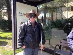 T.J., a male wearing learning shades and holding a long, white cane, waits at a bus stop.