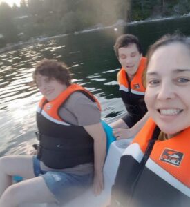 Three people enjoying a day on the water of Deep Bay in a white and blue pedal boat. In the foreground, a woman is smiling at the camera while taking a selfie. She has dark hair pulled back from her face. To her right, in the back of the picture, sits a younger woman, smiling with her eyes closed. She has beautiful curly light brown hair. Behind the latter, a younger man sits slightly smiling and eyes closed. He has brown curly hair. All three of them have light skin and are wearing shorts and black and orange life jackets. The water is calm with a slight ripple, and the late afternoon sun hangs low in the sky. Trees line the far shore, bathed in the soft light of the setting sun.