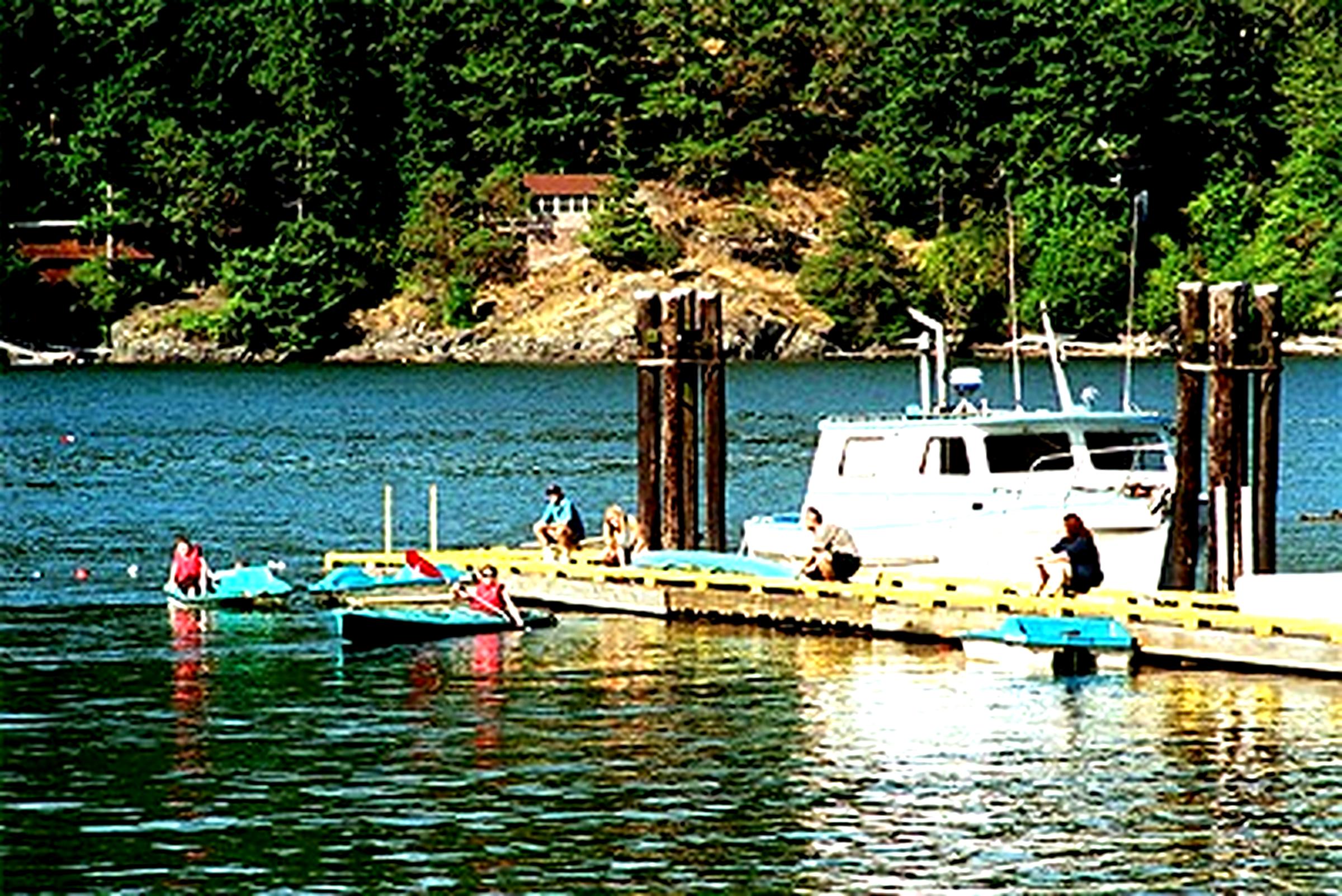 A serene bayside scene on a clear summer day, featuring a dock with several people engaged in leisure activities. Some are sitting on the dock while others paddle small boats. A larger boat is moored at the dock, and the backdrop is a lush, green forested hillside. The overall atmosphere is peaceful and recreational.